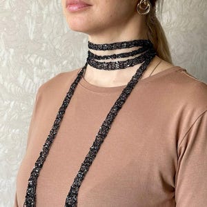 Puede incluir: Un collar de gargantilla negro con cuentas con un diseño largo y colgante. El collar está envuelto alrededor del cuello y cae por la parte delantera. La modelo lleva una camisa marrón y pendientes de aro dorados.