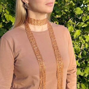 May include: A copper-coloured beaded choker necklace with long, scarf-like strands. The necklace is worn with a matching long-sleeved top and gold hoop earrings. The background features lush green foliage.