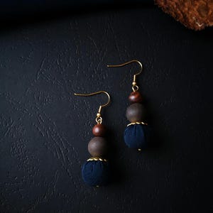 Peut inclure: Une paire de boucles d'oreilles artisanales avec des crochets dorés. Chaque boucle d'oreille est composée d'une perle en bois marron foncé, d'une perle en bois grise et d'une perle recouverte de tissu bleu marine. Design simple et élégant.