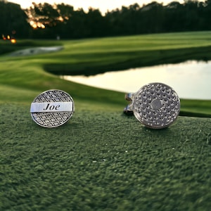 May include: Two silver golf ball markers, one with a textured surface and the other with the name "Joe" engraved on it.