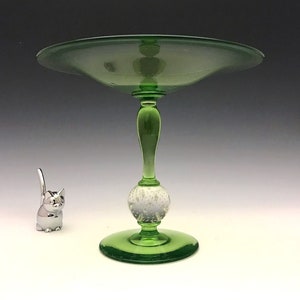 May include: A green glass pedestal bowl with a wide, shallow dish. The stem features a bulbous, textured section and a slender, elongated top. A small, silver cat figurine is to the left of the bowl.