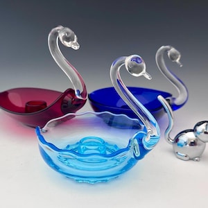 May include: A collection of glass swan-shaped candle holders in shades of blue and red, with clear swan necks. A small, silver cat figurine is also present. The candle holders have a scalloped edge design.