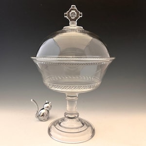 Puede incluir: Una compotera de cristal transparente con tapa abovedada y asa en forma de cruz. La compotera tiene un borde decorativo y se apoya sobre una base de pedestal. Una pequeña figura de gato plateado está junto a la compotera.