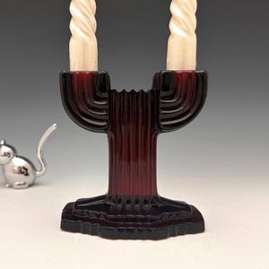 May include: A burgundy glass candelabra with two white spiral candles. The candelabra has a ribbed design and a decorative base. A small silver cat figurine is in the background.