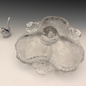 May include: A clear, three-sectioned glass serving dish with a floral pattern and handles. A small, silver cat figurine stands to the left. The dish has a scalloped edge and a central raised section.