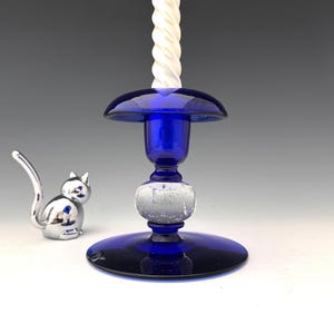 May include: A cobalt blue glass candlestick with a white twisted candle. A silver cat figurine sits to the left. The candlestick has a round base, a bulbous center, and a flared top. The candle is approximately 8 inches tall.