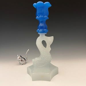 Puede incluir: Una botella de vidrio decorativa con un tapón azul en forma de flor. El cuerpo de la botella tiene forma de pez y se asienta sobre una base en forma de estrella. Una pequeña figura de gato plateado está al lado de la botella.