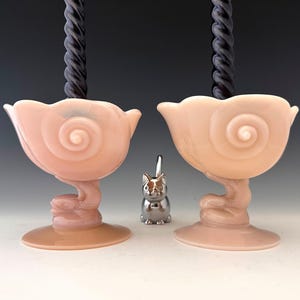 May include: Two pink glass candle holders with a spiral design, a cat-shaped silver-colored object is between them. The candle holders have a seashell-like bowl and a twisted black candle. The base of the candle holders is round.