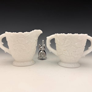 May include: A white milk glass creamer and sugar bowl set with a leaf pattern. The creamer has a spout and handle, while the sugar bowl has two handles. A small silver-colored sugar spoon holder is between the two pieces.