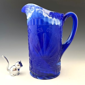May include: A cobalt blue glass pitcher with a handle and spout, featuring the embossed text "GOD BLESS OUR HOME". The pitcher has a decorative design and a white and blue swirled rim. A small, silver cat figurine is next to the pitcher.