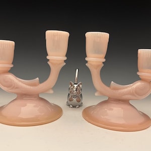 Puede incluir: Dos candelabros de cristal rosa con diseño de pez, cada uno con dos portavelas. Una figura de gato plateado está centrada entre los candelabros. Los candelabros tienen una base redondeada y brazos curvos.