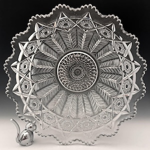 May include: Clear glass serving bowl with a starburst and feather design. The bowl has a scalloped edge and is accompanied by a small, silver cat figurine. The bowl's intricate pattern is visible throughout, creating a decorative piece.