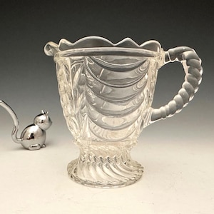 May include: Clear glass creamer with a decorative handle and a scalloped rim. The creamer has a textured pattern and sits on a pedestal base. A small, silver cat figurine is next to the creamer.