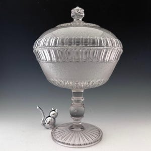 May include: A clear glass candy dish with a lid, featuring a textured surface and a pedestal base. The dish has a round shape with vertical ribbed detailing around the rim. A small silver cat figurine sits beside the dish.