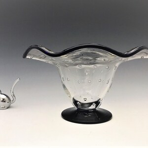 May include: A clear glass bowl with a ruffled rim and a black base. The bowl has small air bubbles throughout. A small, silver cat figurine is to the left of the bowl.