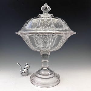 May include: A clear, footed glass candy dish with a lid, featuring intricate embossed patterns. The dish has a wide, scalloped bowl and a decorative finial on the lid. A small, silver cat figurine sits beside the dish.