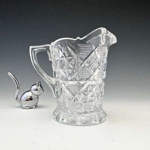 May include: A clear glass pitcher with a diamond pattern and a handle. A small, silver cat figurine is next to the pitcher. The pitcher has a decorative, scalloped rim and a sturdy base. The cat figurine has a long tail.