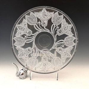 May include: Clear glass plate with a floral design. The plate features an etched pattern of stylized flowers and leaves. A small, silver-colored metal object is positioned near the plate. The plate is displayed on a clear stand.