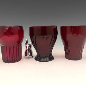 May include: Three ruby red vintage drinking glasses with different designs. The glass on the left has vertical ridges, the middle glass has a spiral design, and the glass on the right has horizontal ridges. A small silver-colored cat figurine is between the glasses.