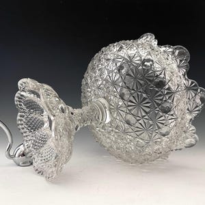 May include: A clear, ornate glass serving dish with a diamond-cut pattern. The dish has a round bowl and a decorative, scalloped edge. It is supported by a clear stem and a silver-colored metal base.