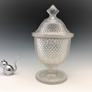 May include: A clear, diamond-patterned glass candy dish with a lid, set on a pedestal base. The dish has a rounded shape and a decorative finial on top. A small, chrome cat figurine sits to the left.