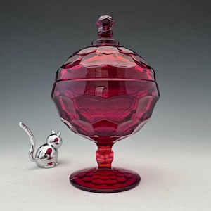 May include: A ruby red, lidded candy dish with a geometric pattern. The dish sits on a pedestal base and has a matching finial on the lid. A small, silver cat figurine is next to the dish.
