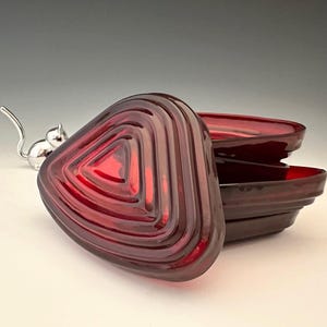 May include: A stack of three triangular, ruby-red glass dishes with a ridged design. A matching lid is in the foreground. A small silver cat figurine is in the background. The dishes have a glossy finish.