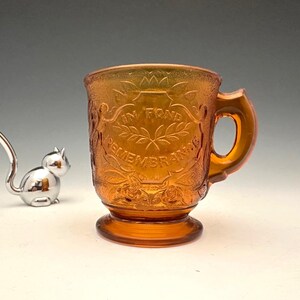 May include: An amber-colored glass mug with a handle and a pedestal base. The mug is embossed with floral and leaf designs and the words "IN FOND REMEMBRANCE". A small silver cat figurine is to the left of the mug.