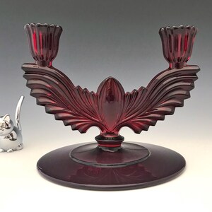 May include: A ruby red glass candelabra with two candle holders. The candelabra has a decorative leaf design and a round base. A silver cat figurine is to the left of the candelabra.