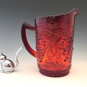 May include: A ruby red glass pitcher with a handle and a decorative grape and leaf pattern. A silver cat figurine is next to the pitcher. The pitcher has a scalloped rim and is likely vintage.