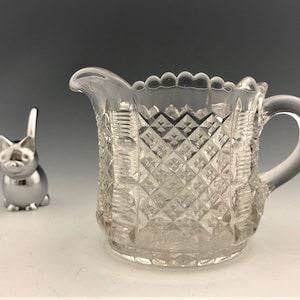 May include: A clear glass creamer with a diamond pattern and a handle. A silver cat-shaped object is to the left of the creamer. The creamer has a scalloped rim. The creamer is likely used for serving cream or milk.
