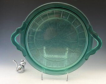 May include: A teal-colored glass serving tray with handles, featuring a detailed floral and geometric pattern. The tray is round and sits on a clear stand. A small silver cat figurine is next to the tray.