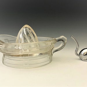 May include: A clear glass citrus juicer with a ribbed reamer and a handle, next to a small, shiny silver cat figurine. The juicer has horizontal and vertical etched lines. The cat figurine has a curved tail and pointed ears.