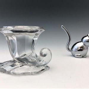 May include: A clear glass horn-shaped vase with a decorative base and a small, silver cat figurine. The vase has a curved design and sits on a detailed base. The cat figurine has a long tail.
