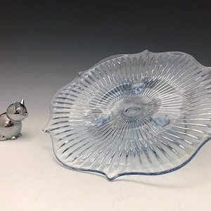 May include: A light blue, scalloped-edge glass serving dish with a ribbed design. Beside it is a silver-colored cat-shaped ring holder. The dish has a raised center and is likely for serving desserts or displaying decorative items.
