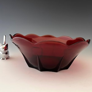 May include: A ruby red glass bowl with a scalloped rim and a silver cat-shaped bottle stopper. The bowl has a faceted design and is set on a white surface. The cat stopper has a red flower detail.