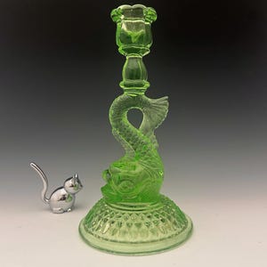 May include: A green glass candlestick holder with a dolphin design. The candlestick has a round base with a diamond pattern and a detailed dolphin figure. A small silver cat figurine is next to the candlestick.