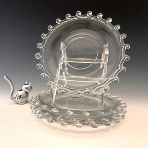 May include: Clear glass plates with a decorative beaded edge are stacked and displayed on a clear stand. A silver cat figurine is positioned to the left of the plates. The plates have a classic, elegant design.