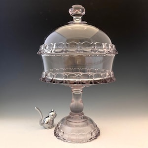 May include: A vintage, light purple glass candy dish with a lid, raised on a pedestal base. The dish has a scalloped edge design and a small, silver cat figurine is next to it.