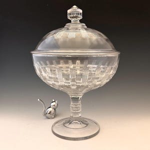 May include: A clear glass candy dish with a lid, featuring a woven basket pattern. The dish sits on a stem and a round base. A small silver cat figurine is next to the dish.