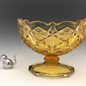 May include: An amber-colored, cut-glass bowl with a pedestal base and a decorative, scalloped rim. The bowl features a diamond and starburst pattern. A small, silver cat figurine is to the left of the bowl.
