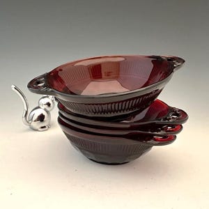 May include: A stack of four ruby red glass bowls with handles. The bowls have a ribbed design and are stacked on top of each other. A silver-colored metal mouse figurine is to the left of the bowls.