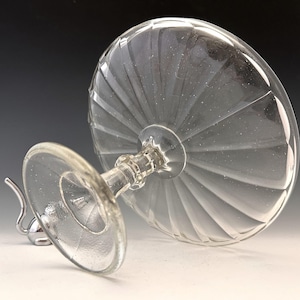May include: Clear glass dessert dish with a pedestal base. The bowl has a ribbed design, and the base is detailed with a faceted pattern. The dish is tilted, showcasing its elegant form and the play of light through the glass.