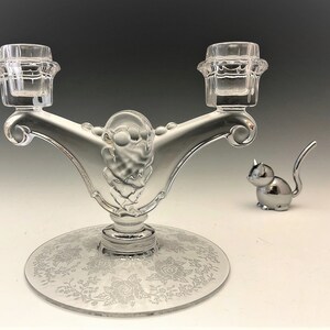 May include: A clear glass candelabra with two candle holders, a decorative floral base, and a silver cat figurine. The candelabra has an ornate design and is made of clear glass.
