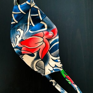 Puede incluir: Una mascarilla con un patrón floral azul, blanco y rojo. La mascarilla tiene dos lazos largos para sujetarla alrededor de la cabeza.