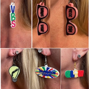 May include: A collection of colorful, novelty earrings. Designs include the text "VBS", "Jesus Shines", "VBS 2026", a lightbulb, a sign that says "The Imagination Station", and the letters "GLOW".