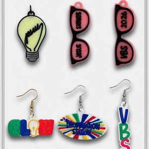 May include: A collection of colorful earrings with religious and inspirational themes. Designs include a lightbulb with "Jesus" written on it, pink glasses with "Jesus Shines", and earrings with the words "Glow", "Illumination Station", and "VBS".