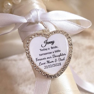 May include: A cream-coloured bridal bouquet with a satin ribbon-wrapped handle. A heart-shaped charm with the name "Jenny" and a message is attached with a white ribbon. The bouquet is decorated with pearls and white satin ribbons.