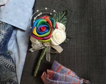 May include: A rainbow rose boutonniere with a white rose and a pearl trim. The boutonniere is pinned to a gray suit jacket with a patterned pocket square.