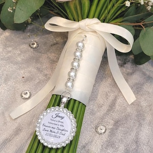 Op de afbeelding: Een zilverkleurige ronde hanger met een rand van strass-steentjes en een wit midden met de tekst: Jenny, Today a Bride, Tomorrow a Wife, Forever our Daughter, Love Mum & Dad, 21/12/2028. De hanger hangt aan een streng witte kunstparels.
